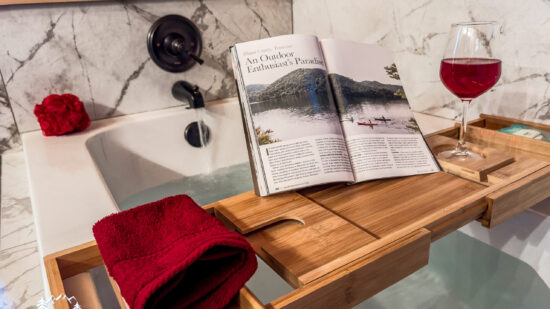A cozy setup with a book, wine glass, and folded towel on a wooden bath tray.